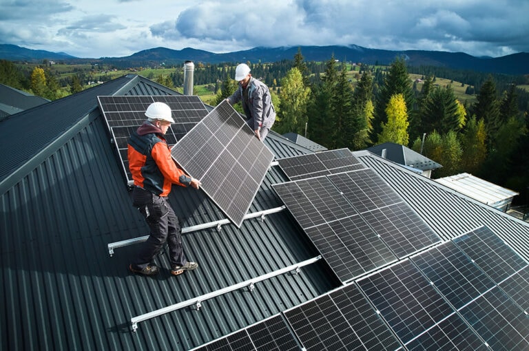 Zwei Personen installieren Solarzellen auf einem Dach, umgeben von bewaldeten Hügeln und bewölktem Himmel.